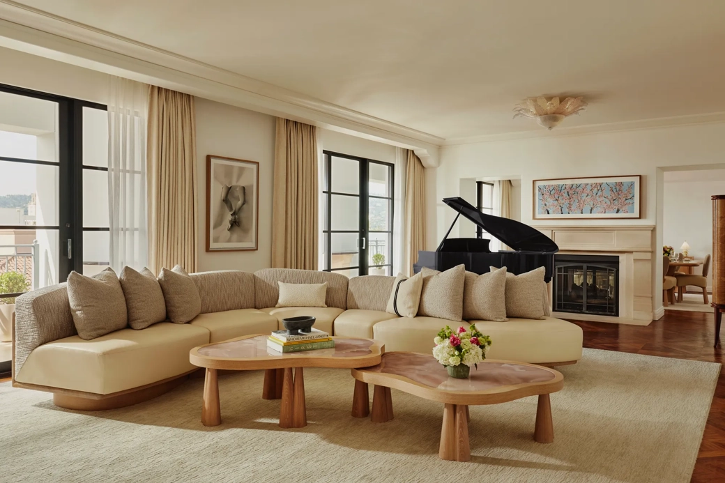 Spacious living room with a curved beige sofa, two wooden coffee tables, large windows with light curtains, a black grand piano, and a fireplace with framed art above.