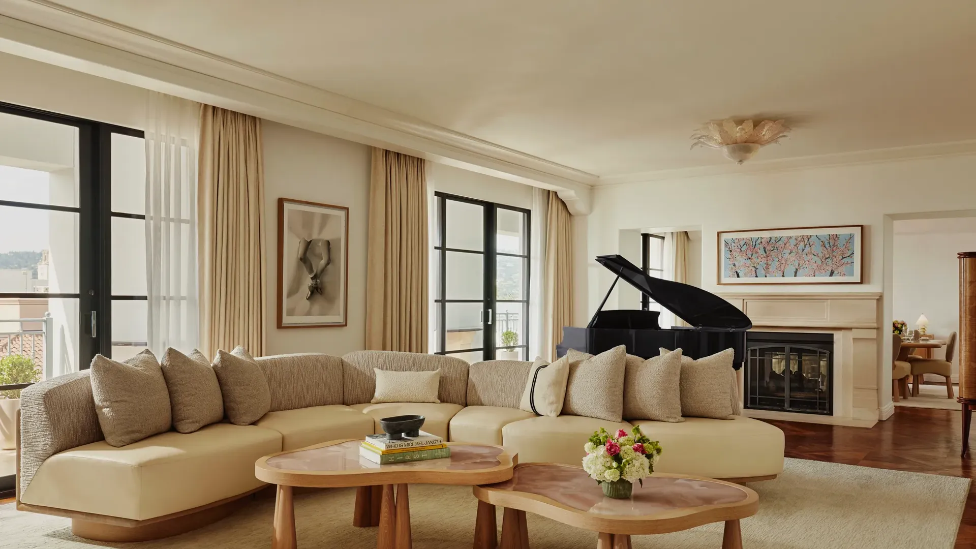 Spacious living room with a curved beige sofa, two wooden coffee tables, large windows with light curtains, a black grand piano, and a fireplace with framed art above.