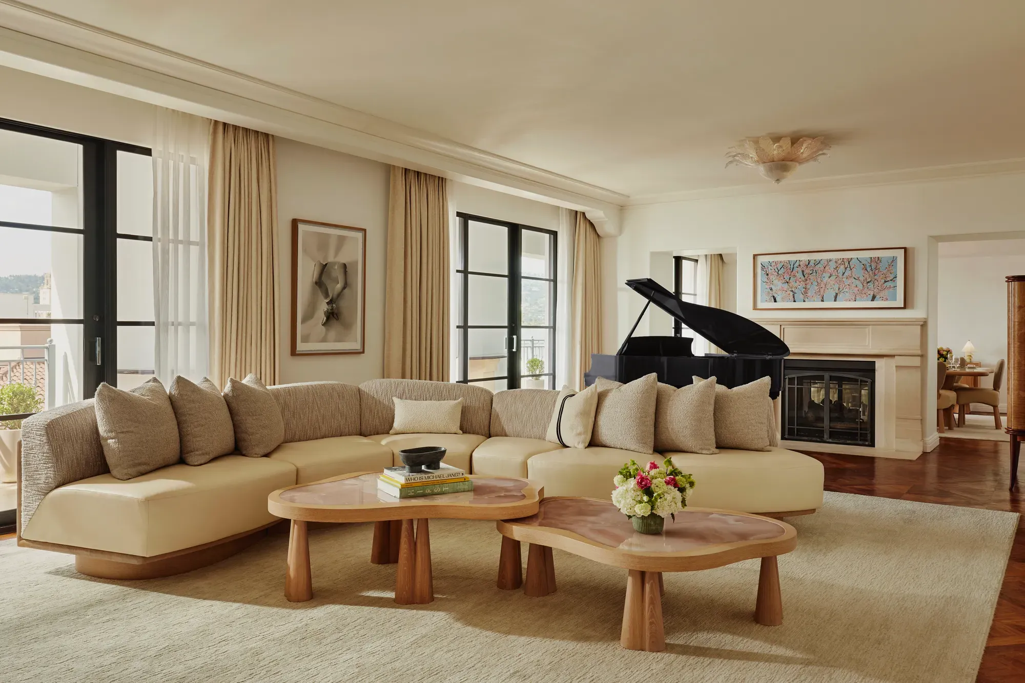 Spacious living room with a curved beige sofa, two wooden coffee tables, large windows with light curtains, a black grand piano, and a fireplace with framed art above.