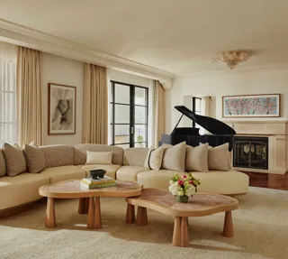 Spacious living room with a curved beige sofa, two wooden coffee tables, large windows with light curtains, a black grand piano, and a fireplace with framed art above.