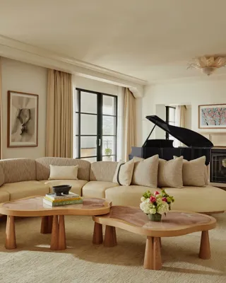 Spacious living room with a curved beige sofa, two wooden coffee tables, large windows with light curtains, a black grand piano, and a fireplace with framed art above.
