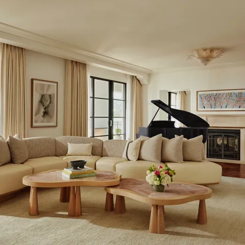 Spacious living room with a curved beige sofa, two wooden coffee tables, large windows with light curtains, a black grand piano, and a fireplace with framed art above.