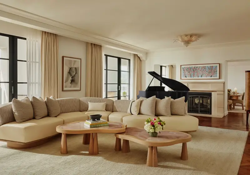 Spacious living room with a curved beige sofa, two wooden coffee tables, large windows with light curtains, a black grand piano, and a fireplace with framed art above.