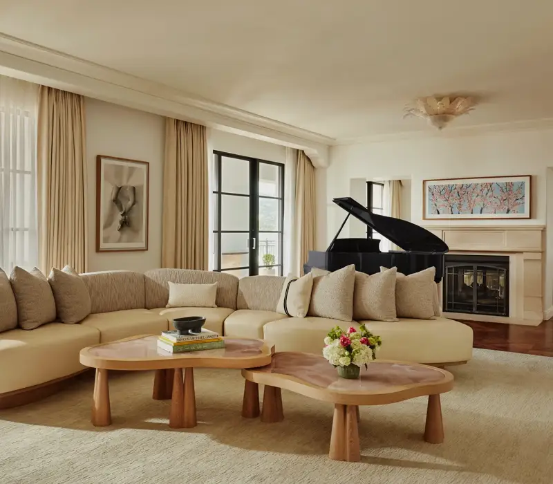 Spacious living room with a curved beige sofa, two wooden coffee tables, large windows with light curtains, a black grand piano, and a fireplace with framed art above.