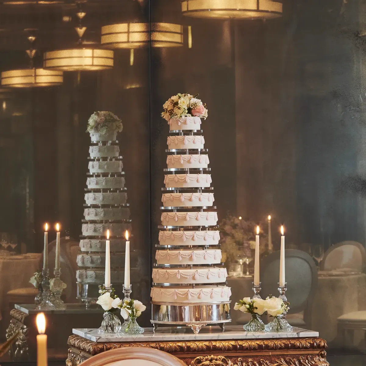 Tall tiered wedding cake displayed on ornate table with candles and floral accents.