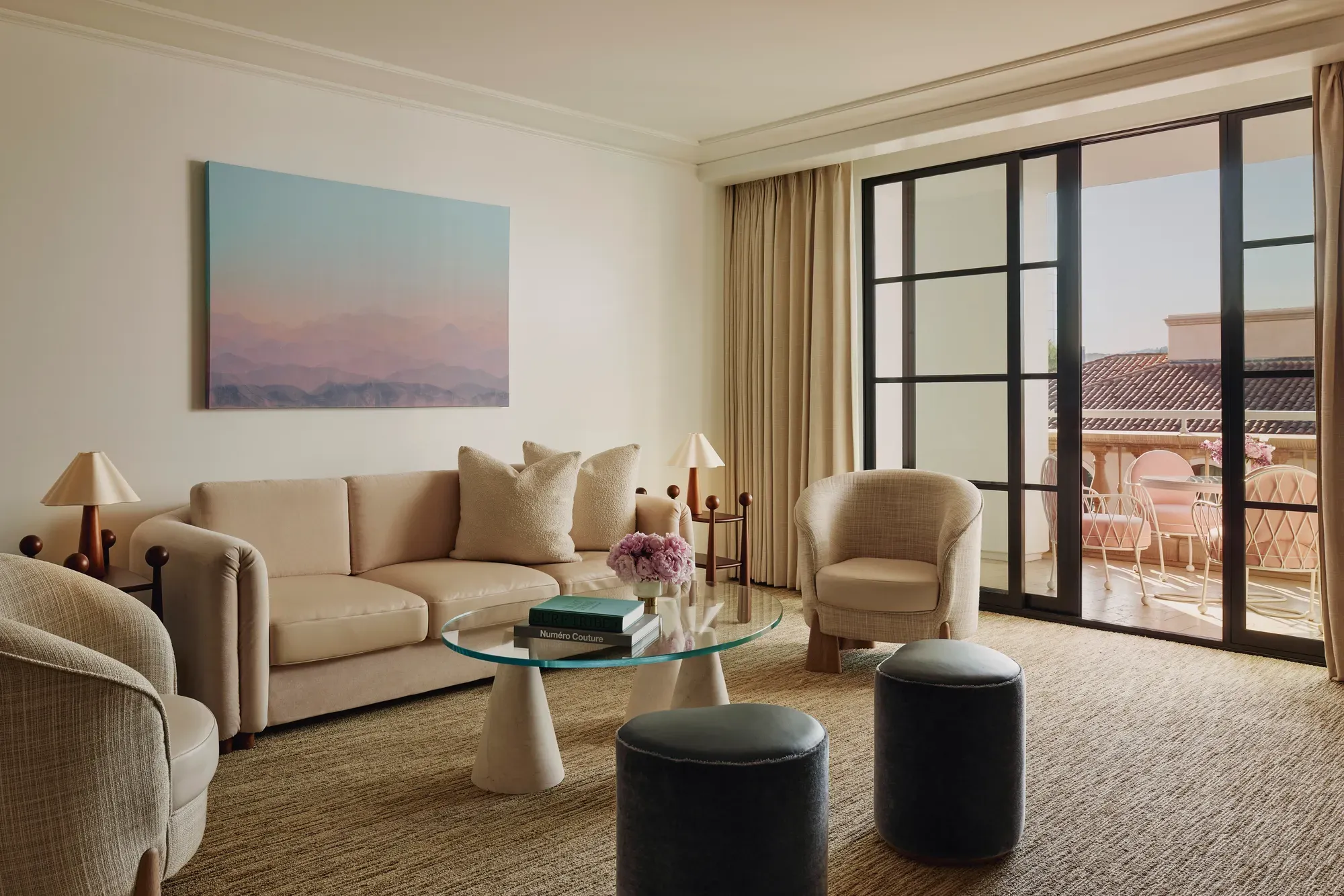 Living room with cream sofa, two armchairs, glass coffee table and ottomans, large landscape artwork, and doors to a terrace with seating.