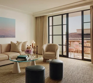 Living room with cream sofa, two armchairs, glass coffee table and ottomans, large landscape artwork, and doors to a terrace with seating.