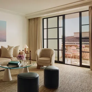 Living room with cream sofa, two armchairs, glass coffee table and ottomans, large landscape artwork, and doors to a terrace with seating.