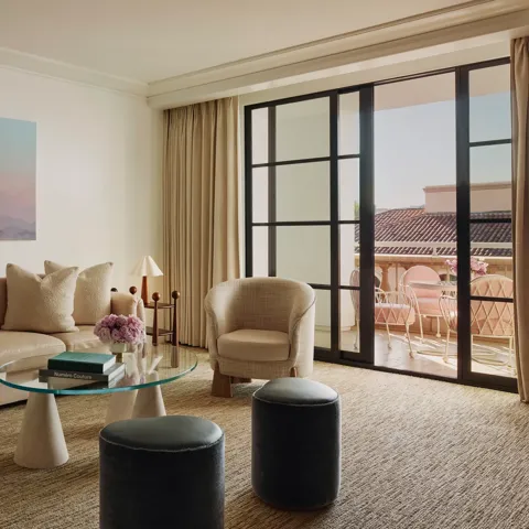 Living room with cream sofa, two armchairs, glass coffee table and ottomans, large landscape artwork, and doors to a terrace with seating.