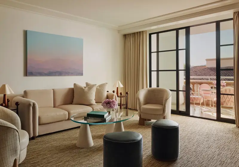 Living room with cream sofa, two armchairs, glass coffee table and ottomans, large landscape artwork, and doors to a terrace with seating.