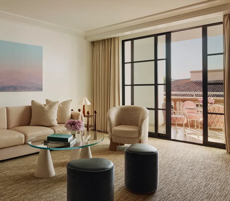 Living room with cream sofa, two armchairs, glass coffee table and ottomans, large landscape artwork, and doors to a terrace with seating.