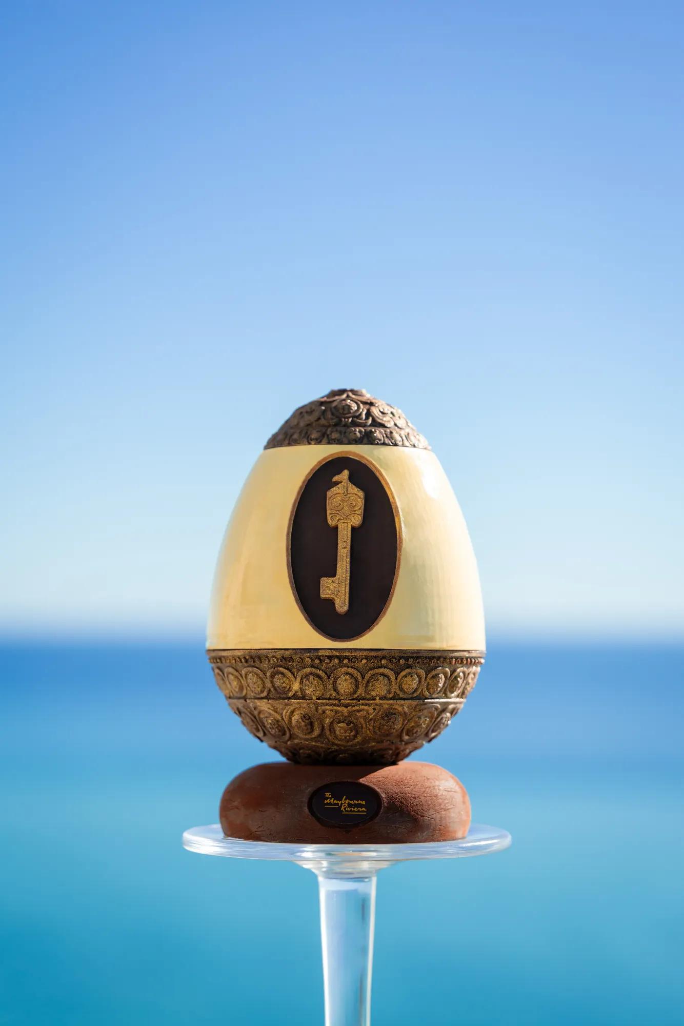 Sculpture d’œuf décoratif avec base ornée et motif de clé, présentée sur un support en verre face à la mer bleue.