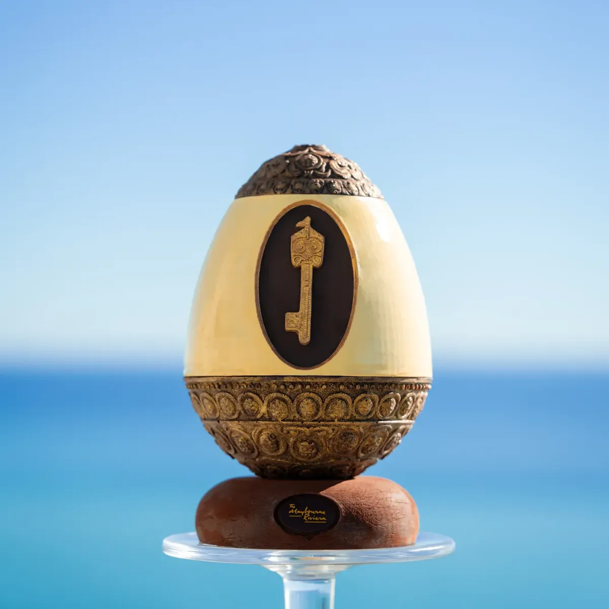 Sculpture d’œuf décoratif avec base ornée et motif de clé, présentée sur un support en verre face à la mer bleue.