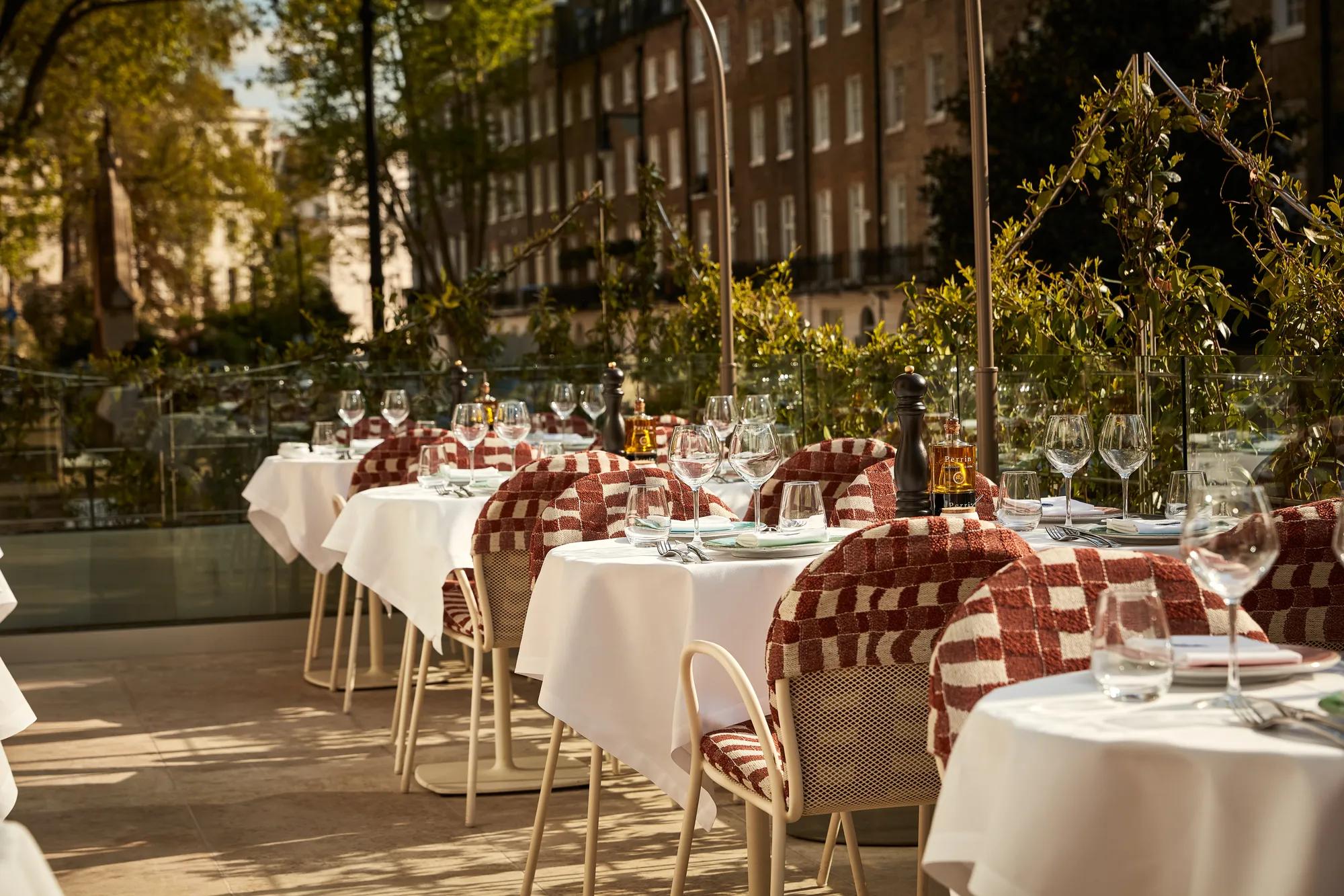 Outdoor terrace dining area with white tablecloths, red patterned chairs, and glassware in afternoon sunlight.