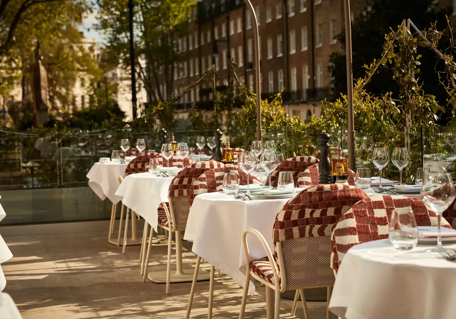 Outdoor terrace dining area with white tablecloths, red patterned chairs, and glassware in afternoon sunlight.