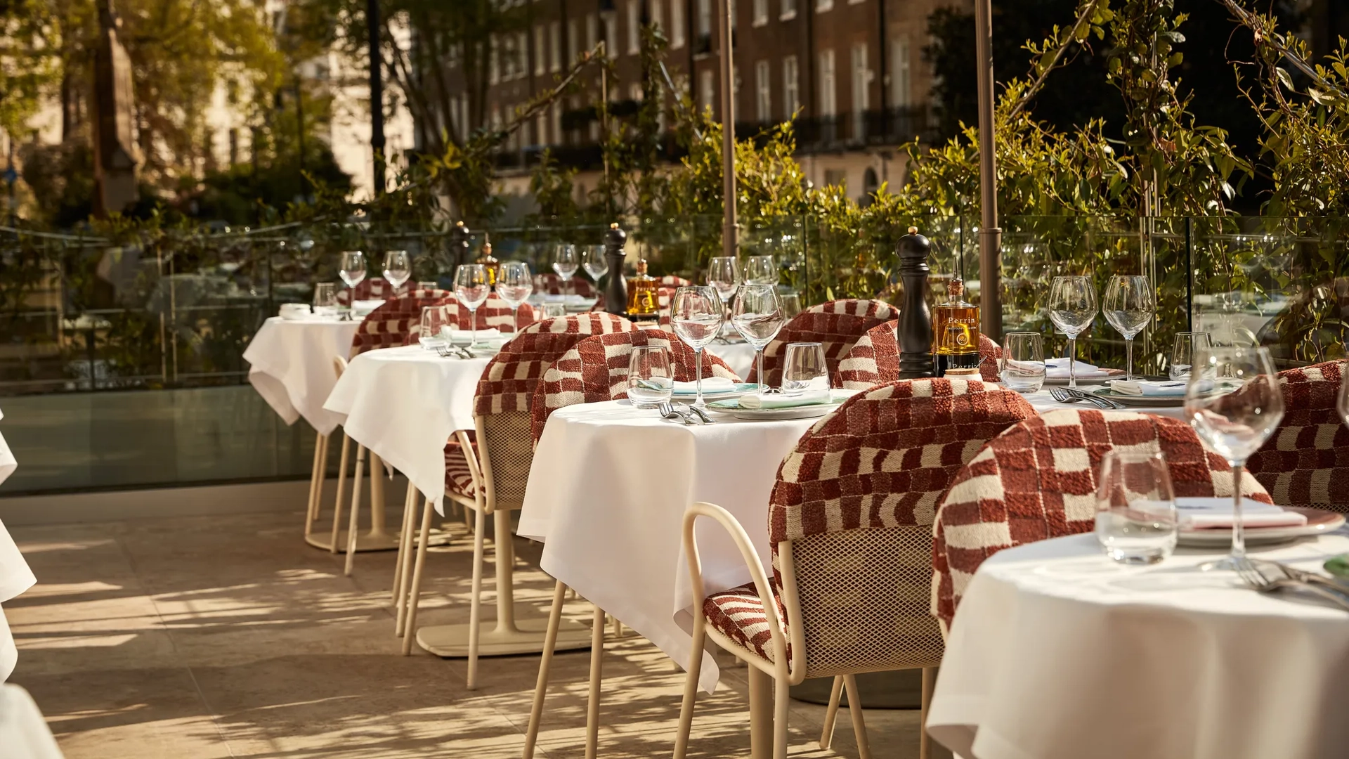 Outdoor terrace dining area with white tablecloths, red patterned chairs, and glassware in afternoon sunlight.