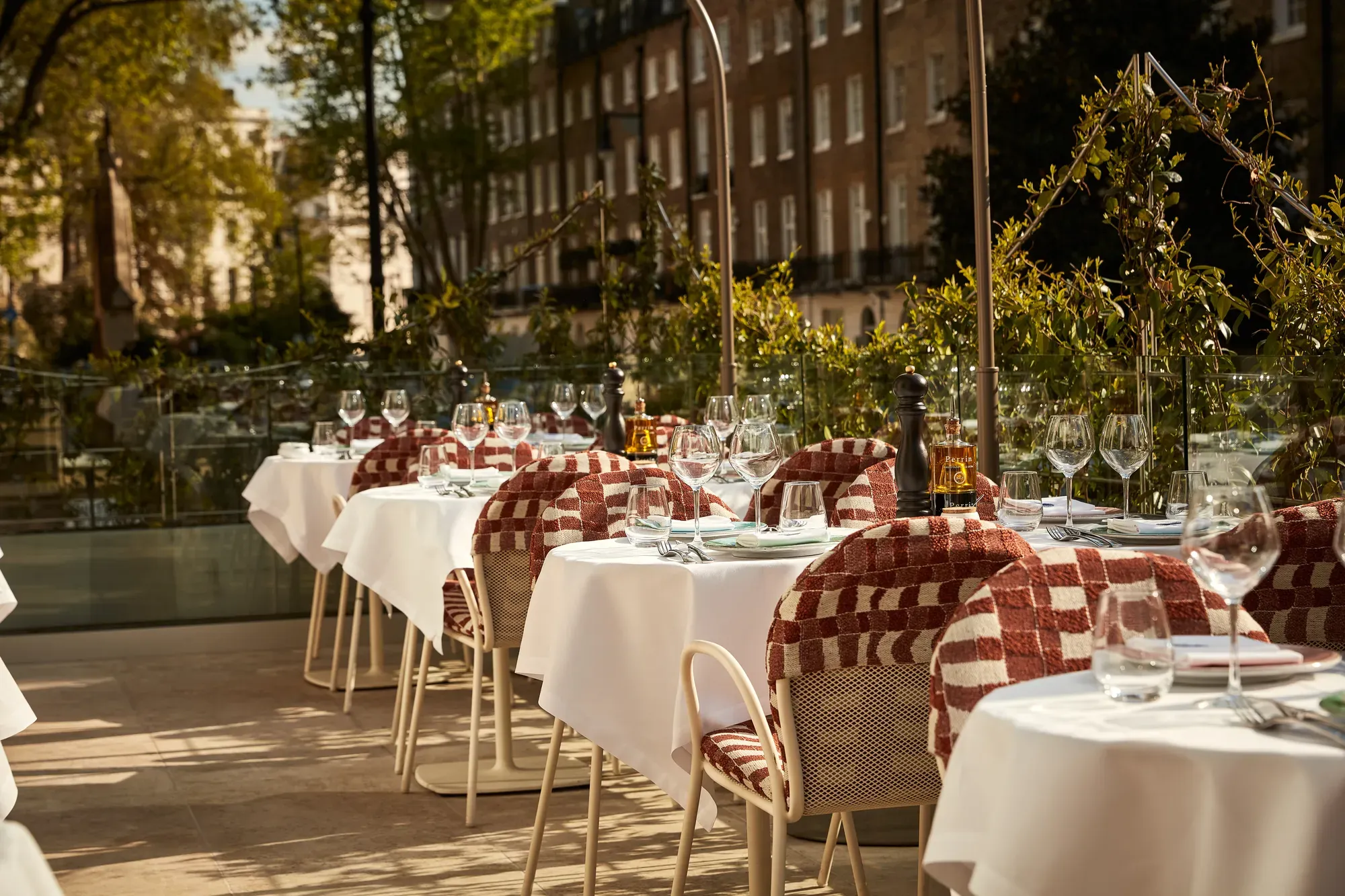 Outdoor terrace dining area with white tablecloths, red patterned chairs, and glassware in afternoon sunlight.