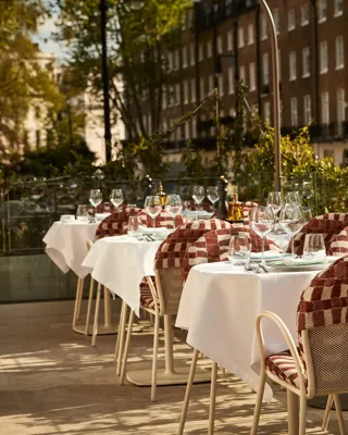 Outdoor terrace dining area with white tablecloths, red patterned chairs, and glassware in afternoon sunlight.