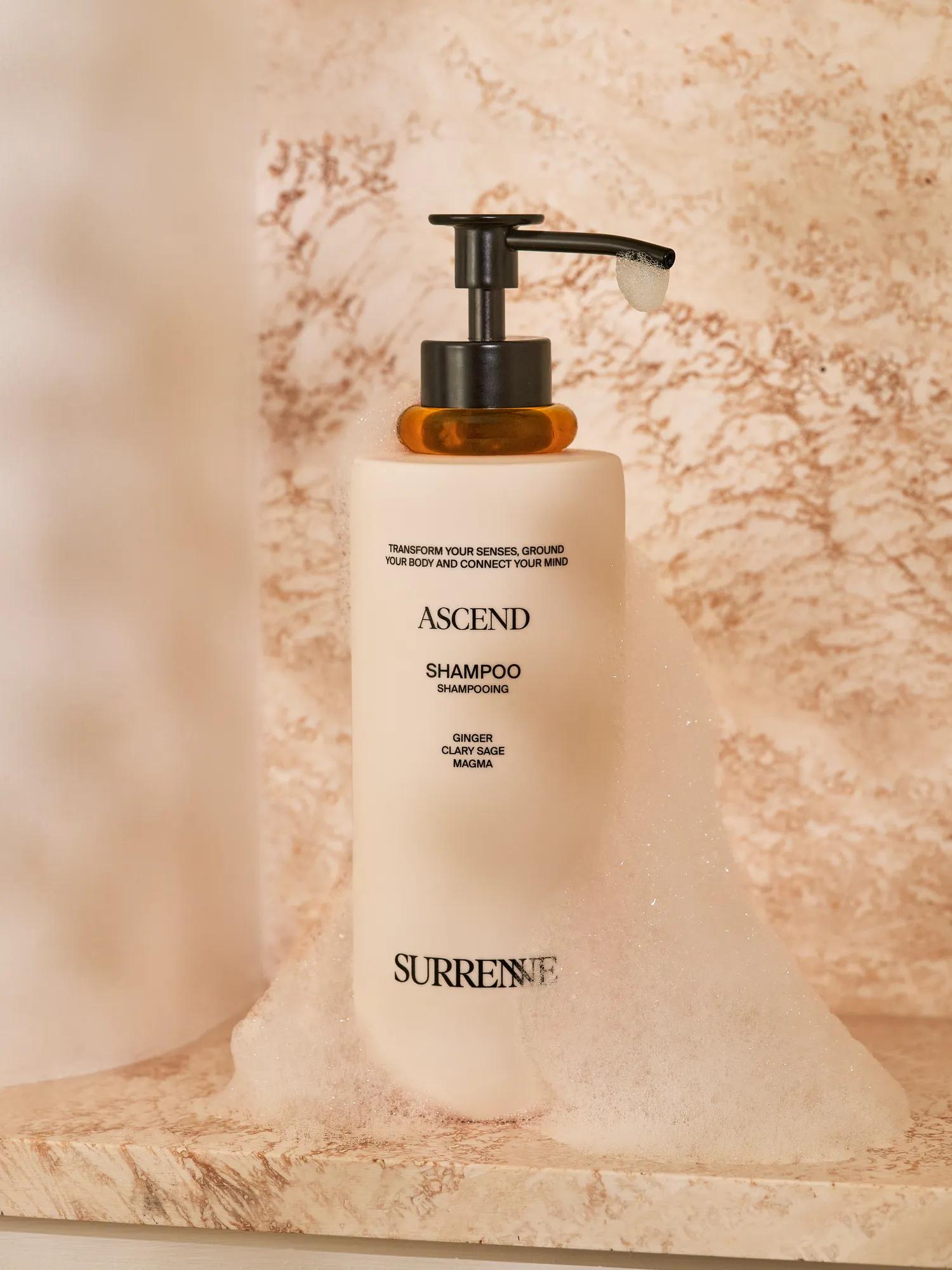 Surrenne Ascend shampoo bottle surrounded by foam on a marble shower shelf with warm ambient lighting.