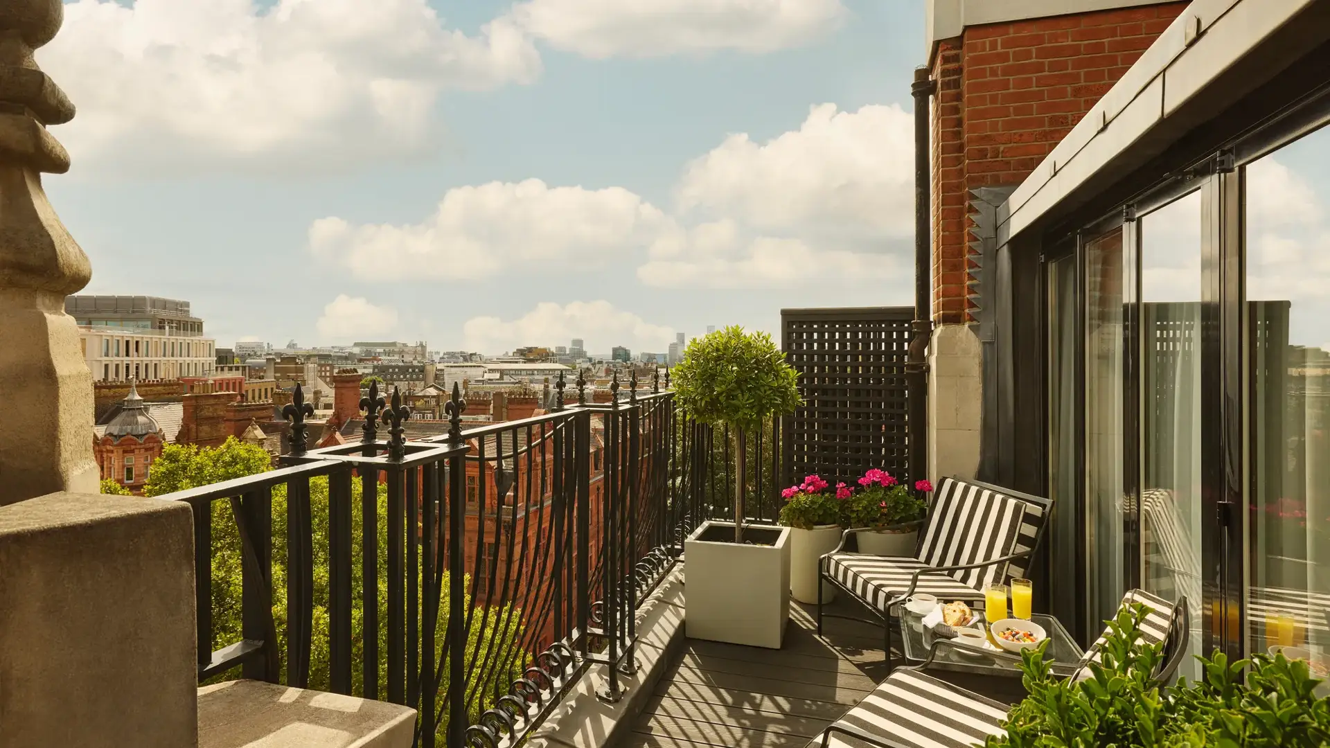 Private balcony with striped loungers, potted plants, and city skyline views under a blue sky.