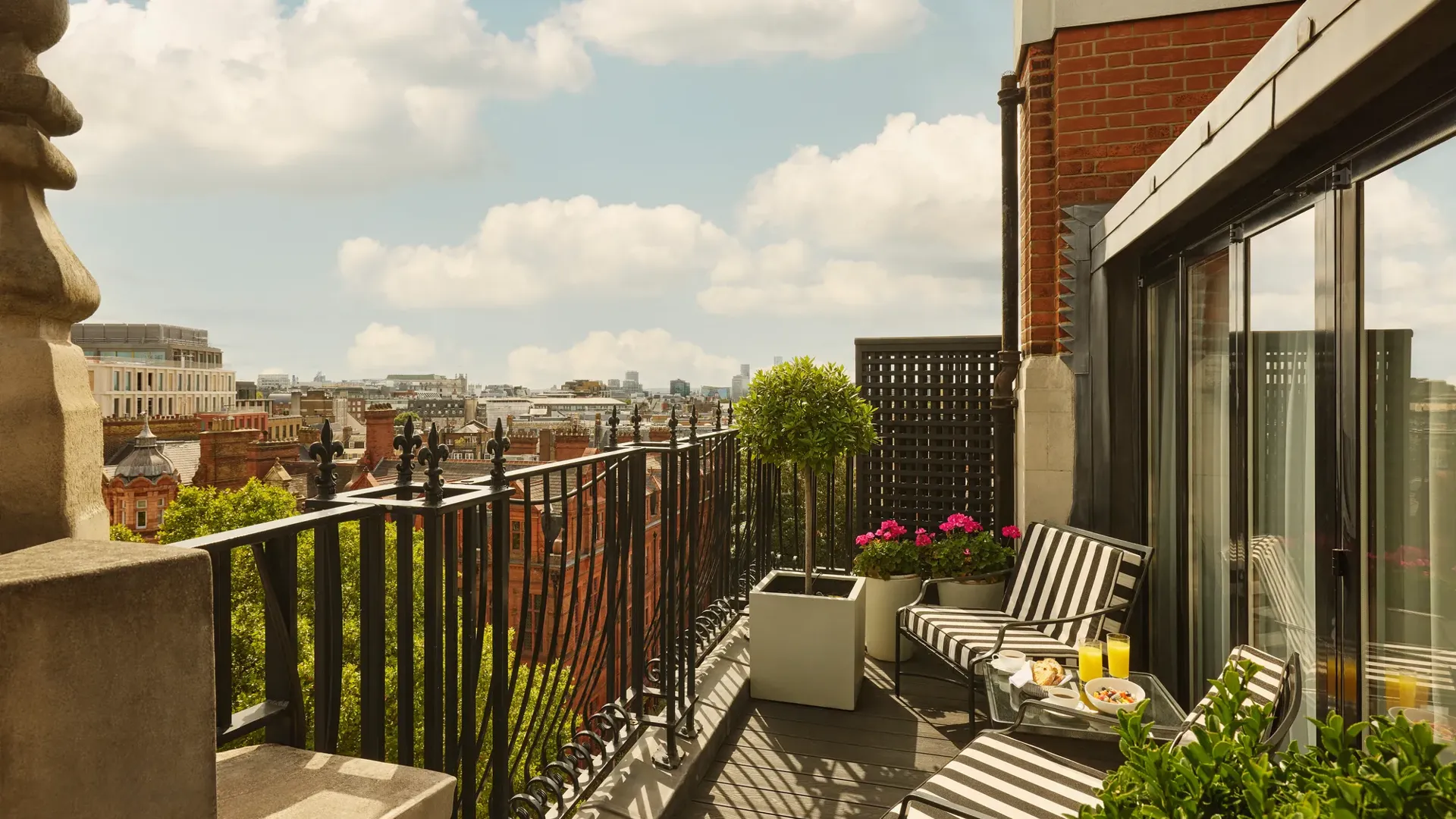 Private balcony with striped loungers, potted plants, and city skyline views under a blue sky.