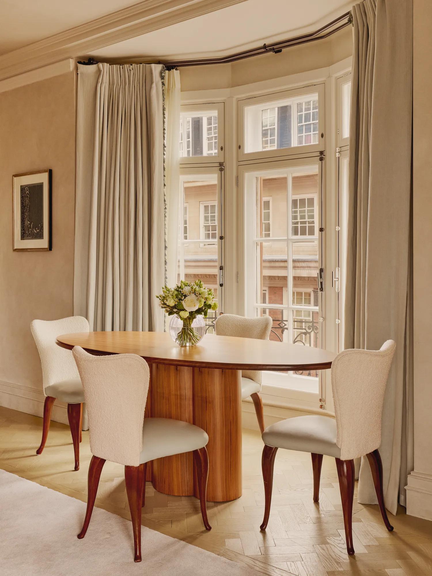 Bright dining area with a round wooden table and four cream upholstered chairs set beside tall bay windows framed with soft beige curtains and a vase of fresh flowers as the centerpiece.