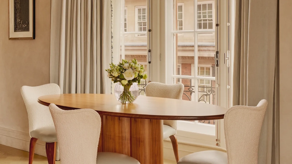 Bright dining area with a round wooden table and four cream upholstered chairs set beside tall bay windows framed with soft beige curtains and a vase of fresh flowers as the centerpiece.