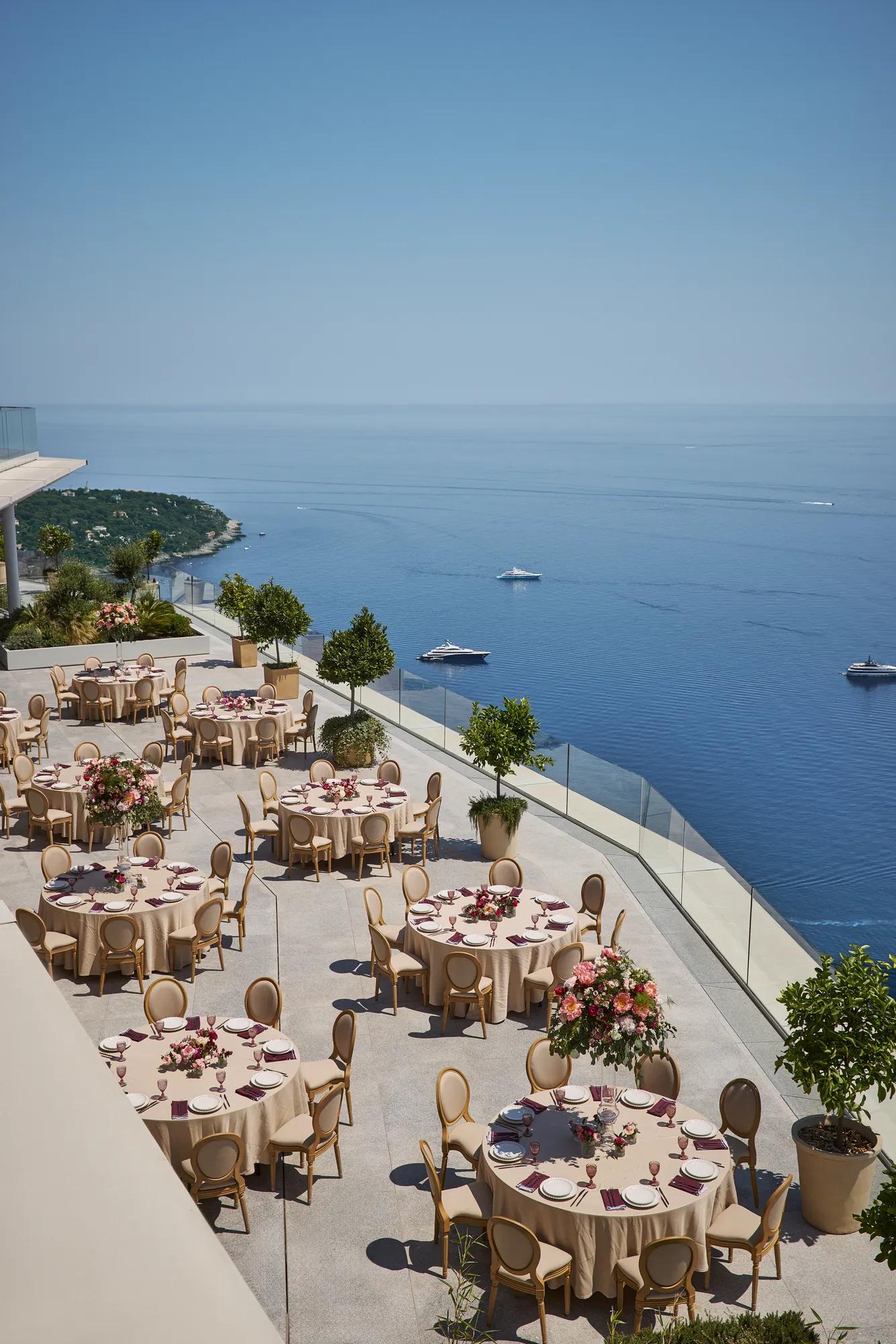Terrace set for an outdoor event with round tables covered in beige cloths, decorated with pink floral centerpieces and elegant chairs, overlooking the sea with yachts in the distance.