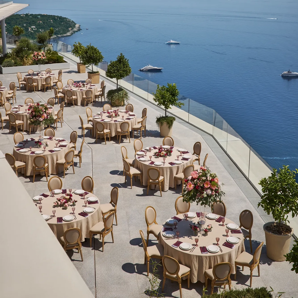 Terrace set for an outdoor event with round tables covered in beige cloths, decorated with pink floral centerpieces and elegant chairs, overlooking the sea with yachts in the distance.