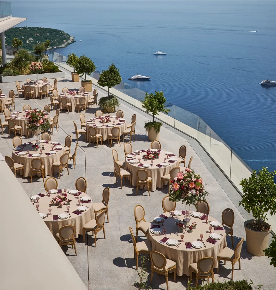Terrace set for an outdoor event with round tables covered in beige cloths, decorated with pink floral centerpieces and elegant chairs, overlooking the sea with yachts in the distance.