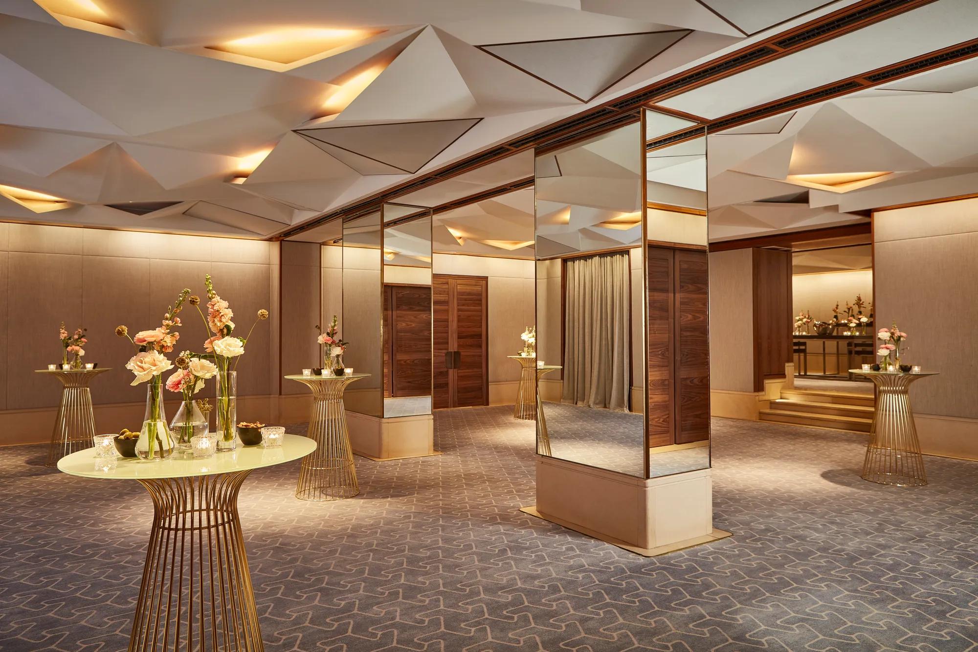 Elegant pre-function space with mirrored columns, gold tables, and floral decorations under geometric ceiling lights.