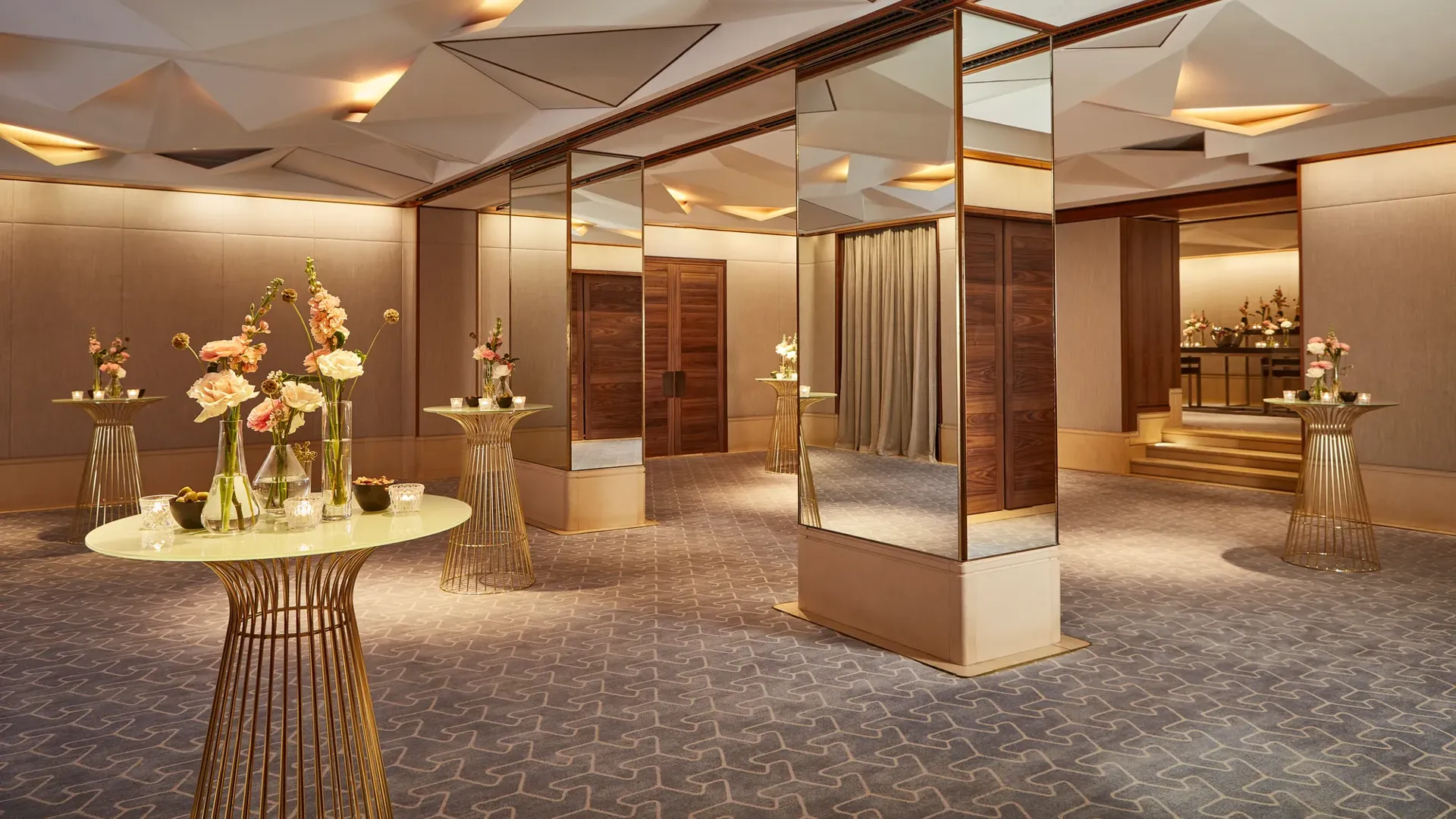 Elegant pre-function space with mirrored columns, gold tables, and floral decorations under geometric ceiling lights.