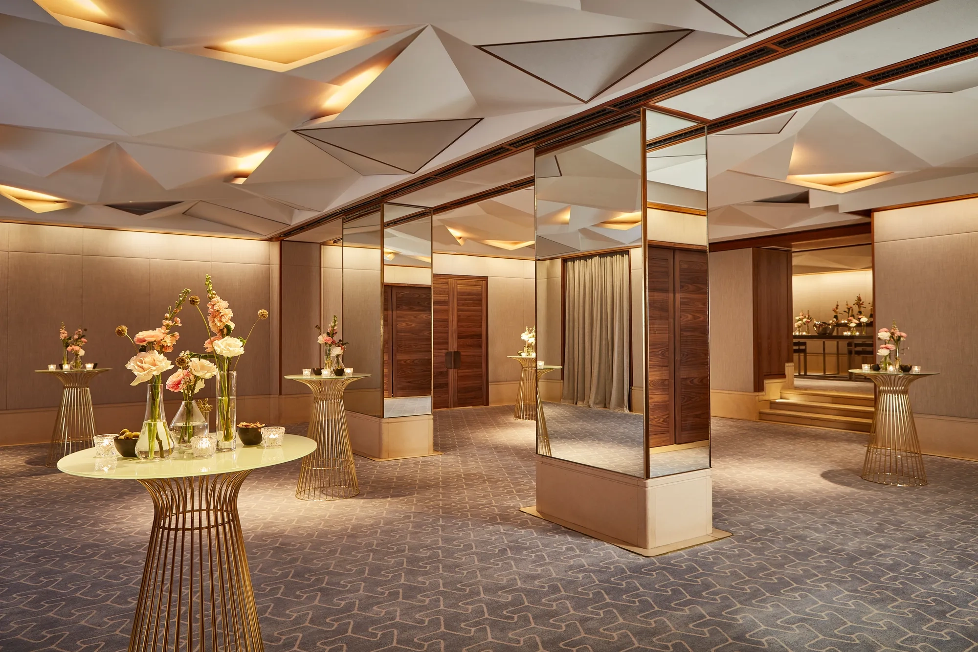 Elegant pre-function space with mirrored columns, gold tables, and floral decorations under geometric ceiling lights.