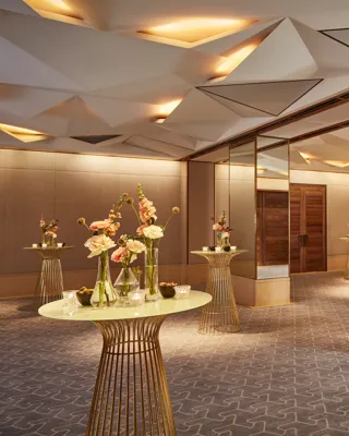 Elegant pre-function space with mirrored columns, gold tables, and floral decorations under geometric ceiling lights.