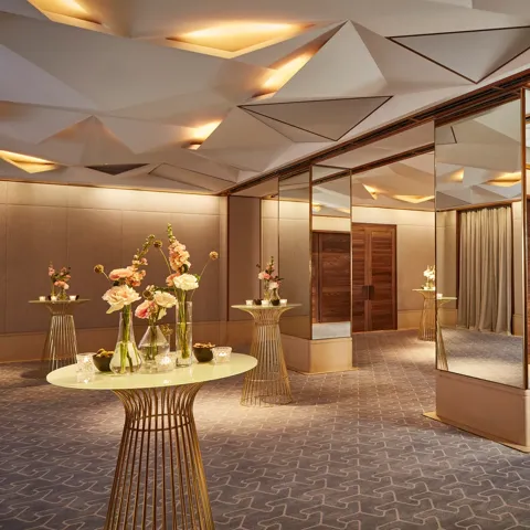 Elegant pre-function space with mirrored columns, gold tables, and floral decorations under geometric ceiling lights.