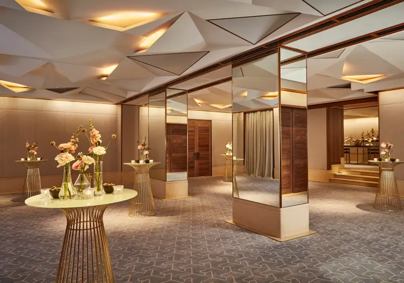 Elegant pre-function space with mirrored columns, gold tables, and floral decorations under geometric ceiling lights.
