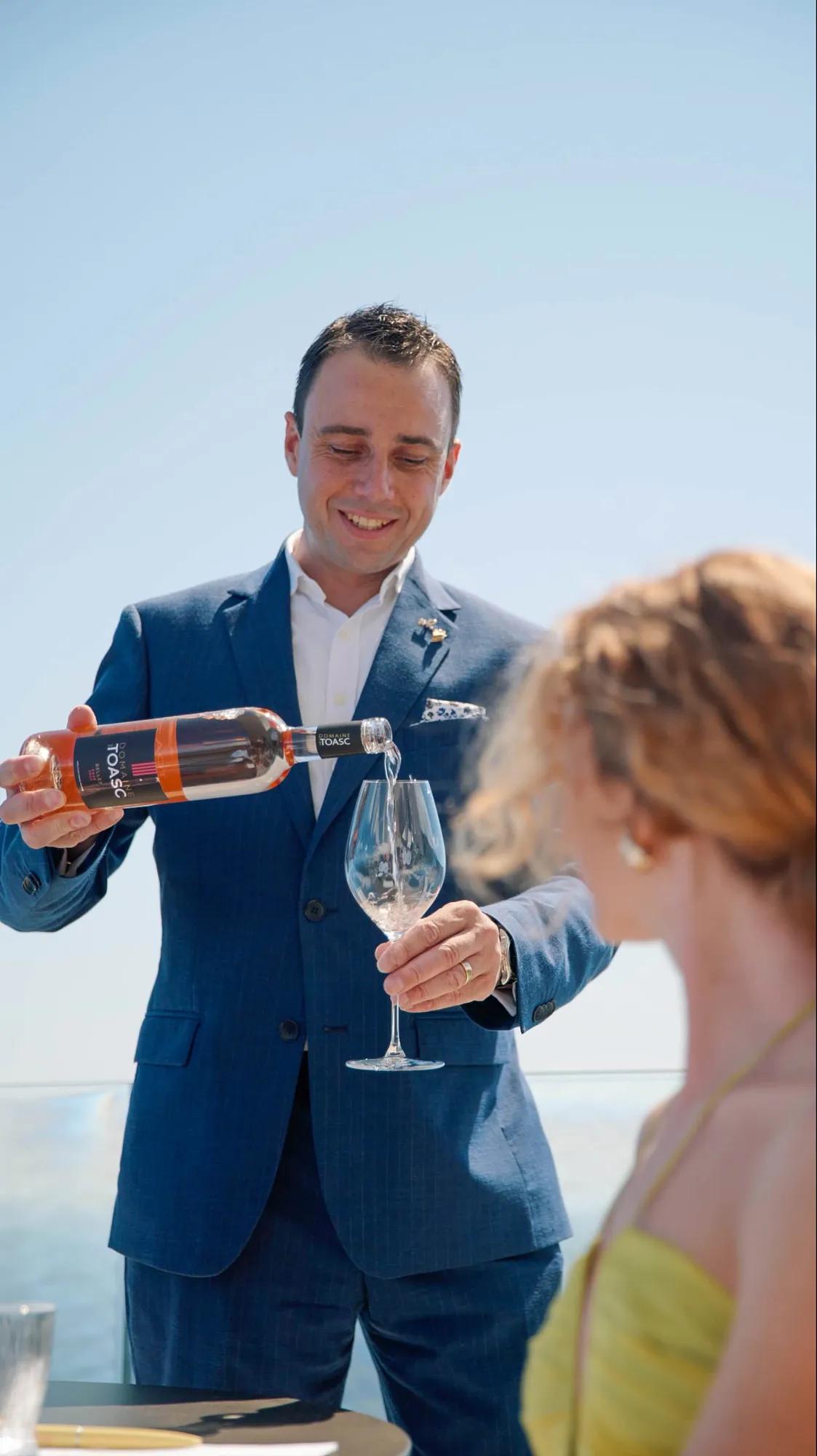 Serveur en costume bleu versant du rosé dans le verre d’une cliente sur une terrasse ensoleillée.