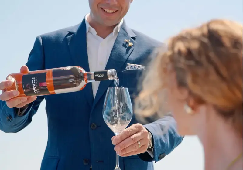 Serveur en costume bleu versant du rosé dans le verre d’une cliente sur une terrasse ensoleillée.