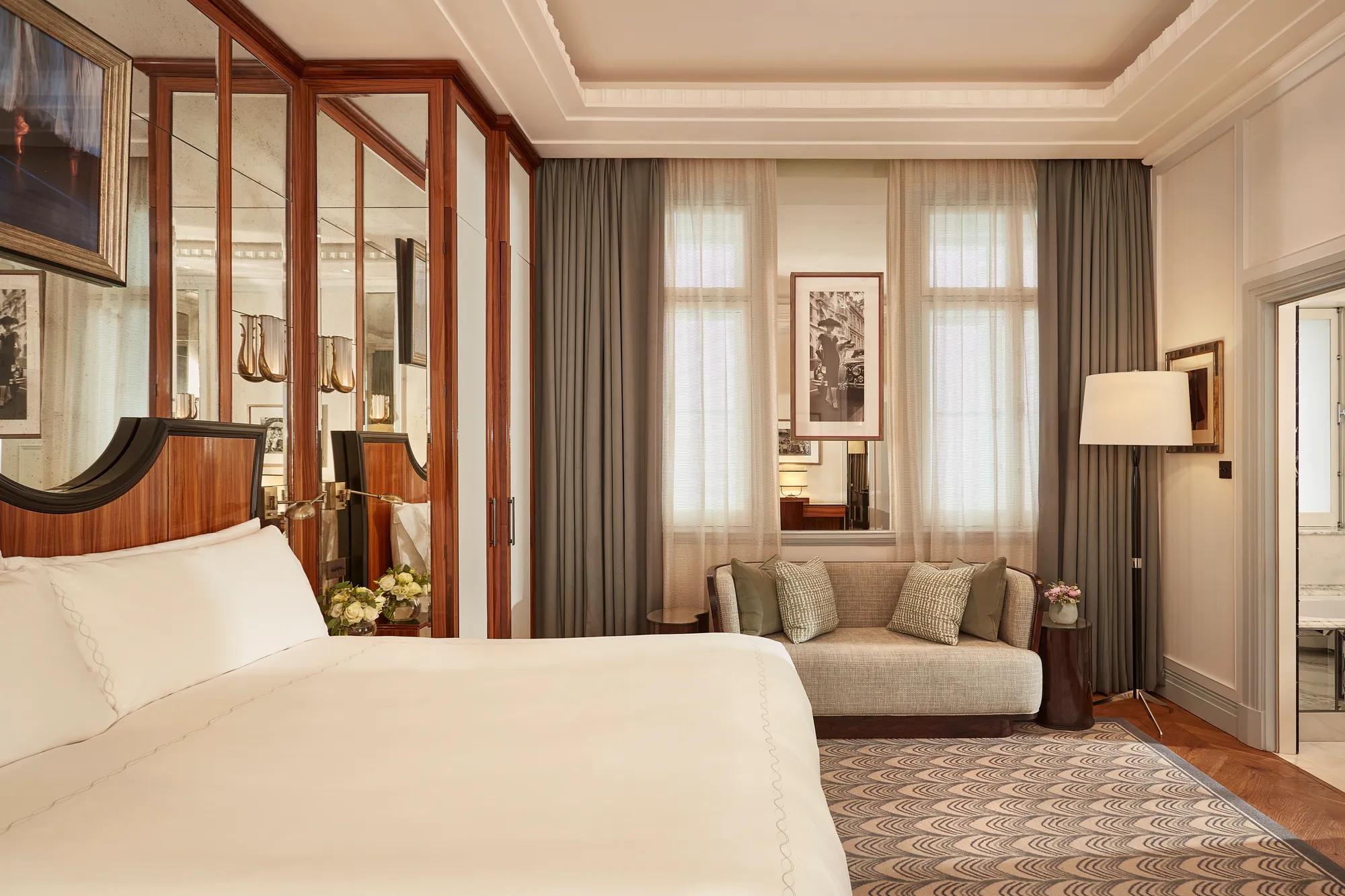 Refined bedroom with a king-size bed, mirrored wardrobe doors, small sofa beneath tall windows with sheer curtains, and framed artwork on the walls.