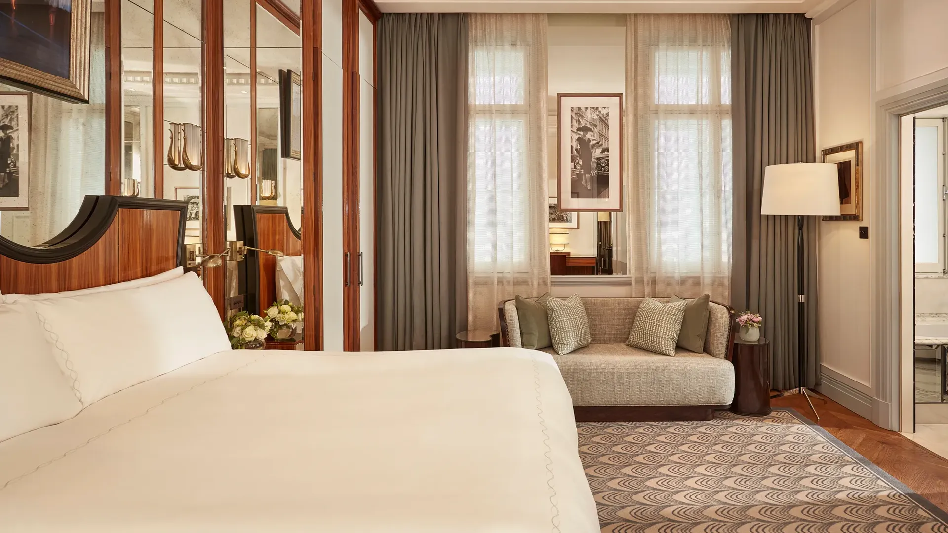 Refined bedroom with a king-size bed, mirrored wardrobe doors, small sofa beneath tall windows with sheer curtains, and framed artwork on the walls.