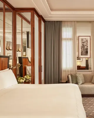 Refined bedroom with a king-size bed, mirrored wardrobe doors, small sofa beneath tall windows with sheer curtains, and framed artwork on the walls.
