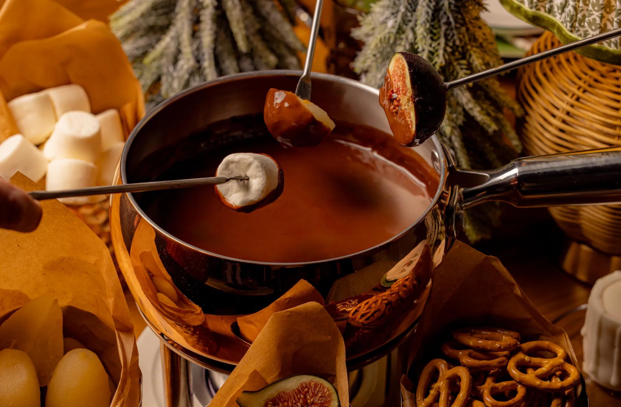 Chocolate fondue pot with skewered fruit and marshmallows, surrounded by festive treats and winter foliage.