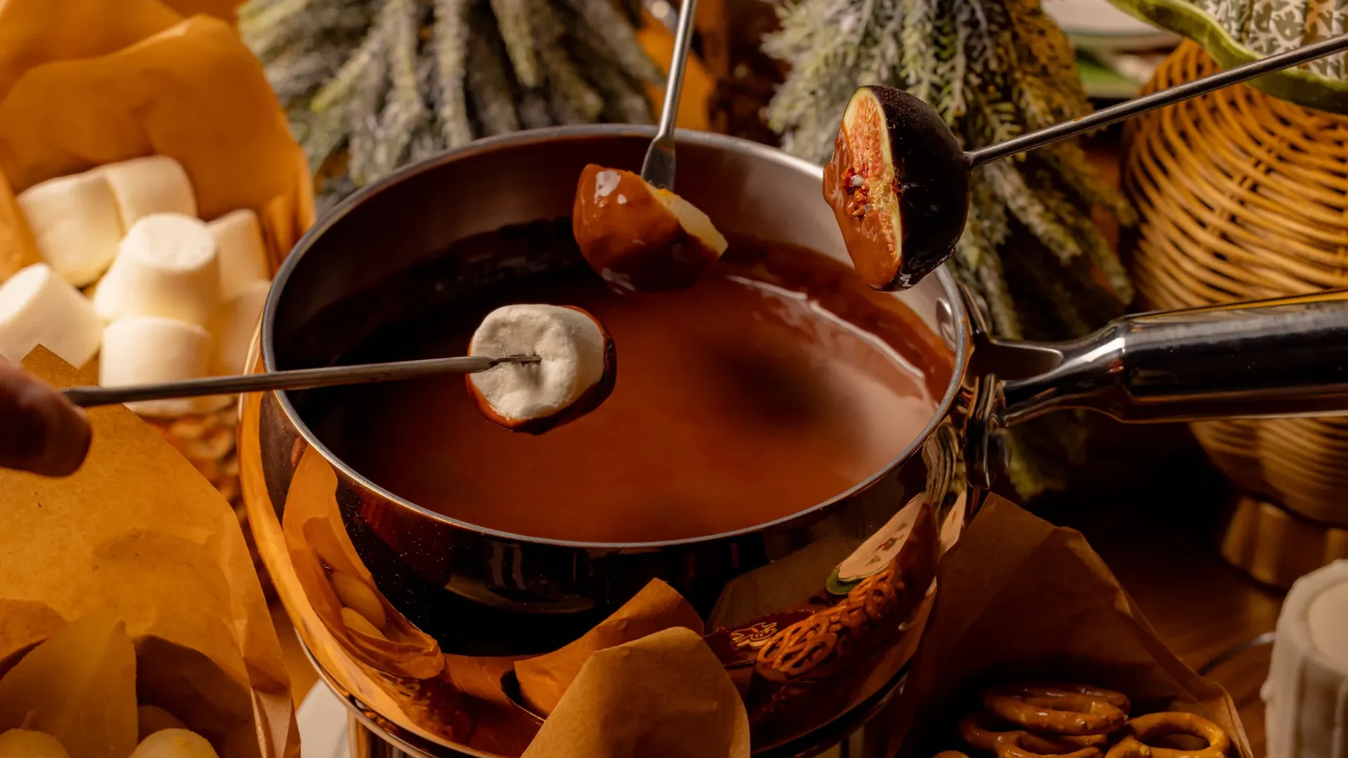 Chocolate fondue pot with skewered fruit and marshmallows, surrounded by festive treats and winter foliage.