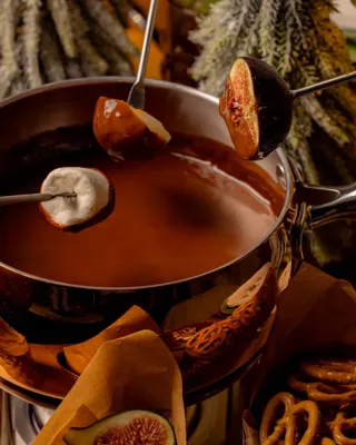 Chocolate fondue pot with skewered fruit and marshmallows, surrounded by festive treats and winter foliage.