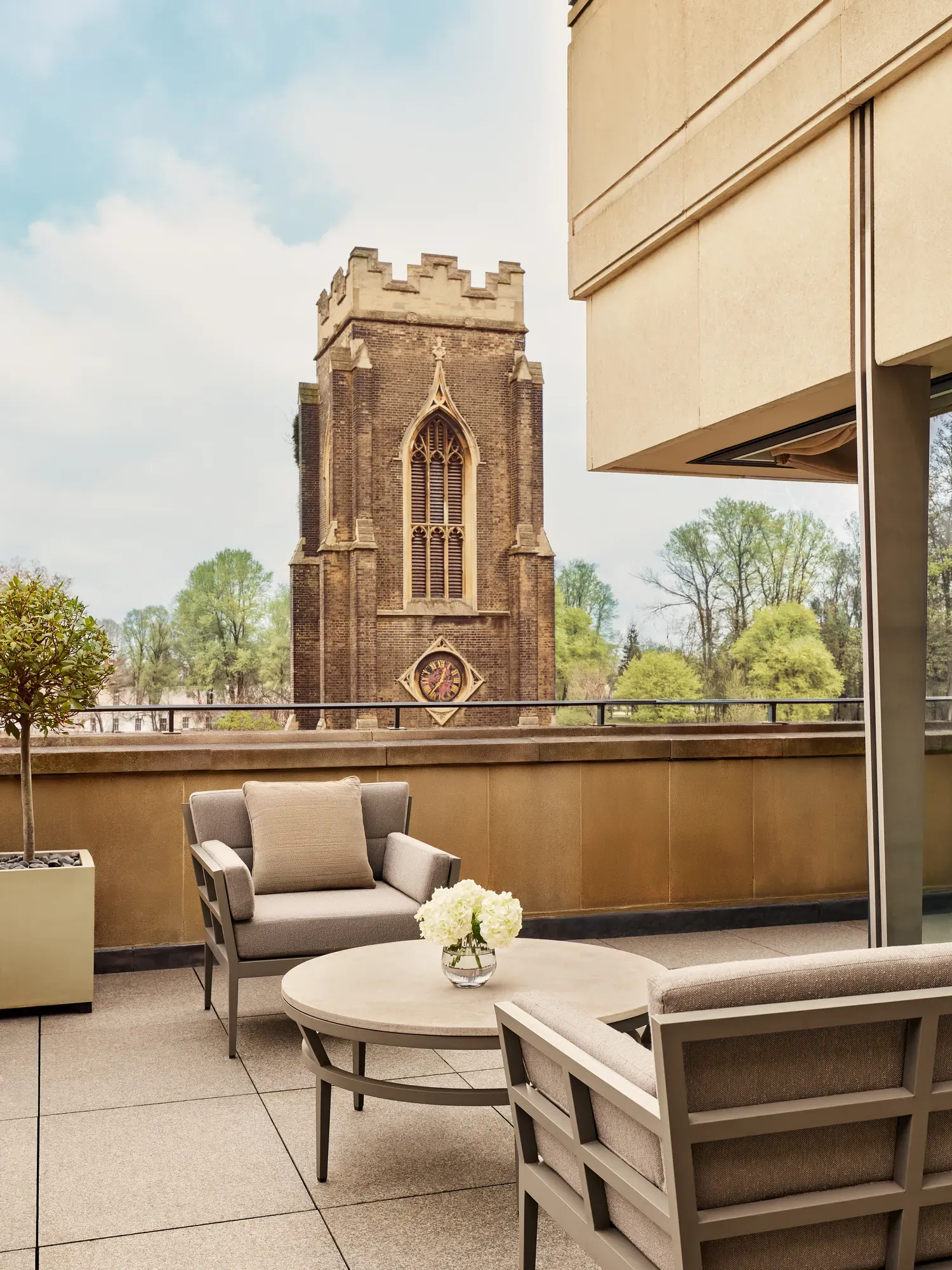 A rooftop terrace with cushioned chairs and a round table decorated with white flowers overlooks a historic stone clocktower surrounded by green trees under a bright sky.