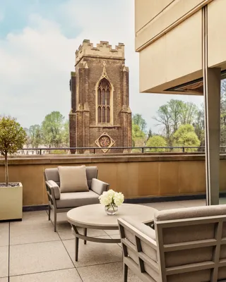 A rooftop terrace with cushioned chairs and a round table decorated with white flowers overlooks a historic stone clocktower surrounded by green trees under a bright sky.