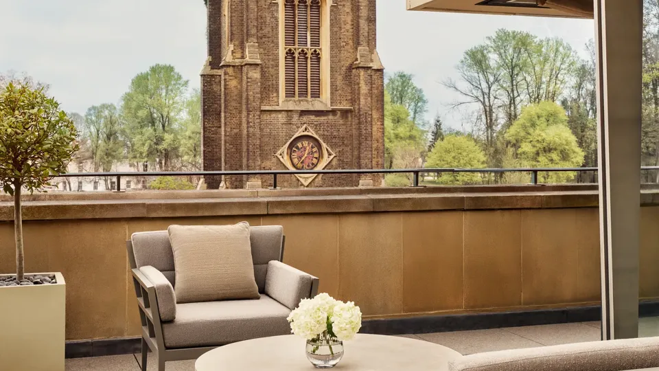 A rooftop terrace with cushioned chairs and a round table decorated with white flowers overlooks a historic stone clocktower surrounded by green trees under a bright sky.