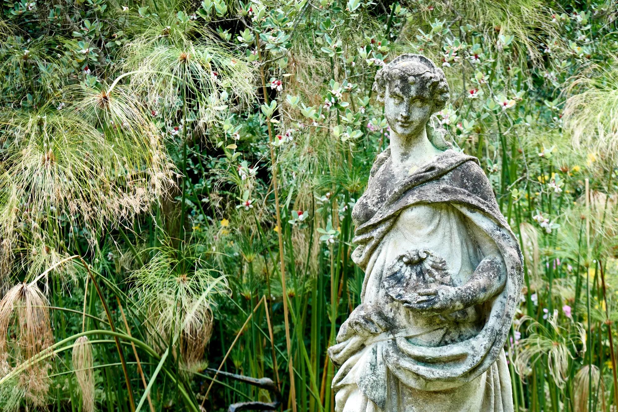 Statue en pierre patinée représentant une femme tenant un coquillage, entourée de hautes herbes et de fleurs.
