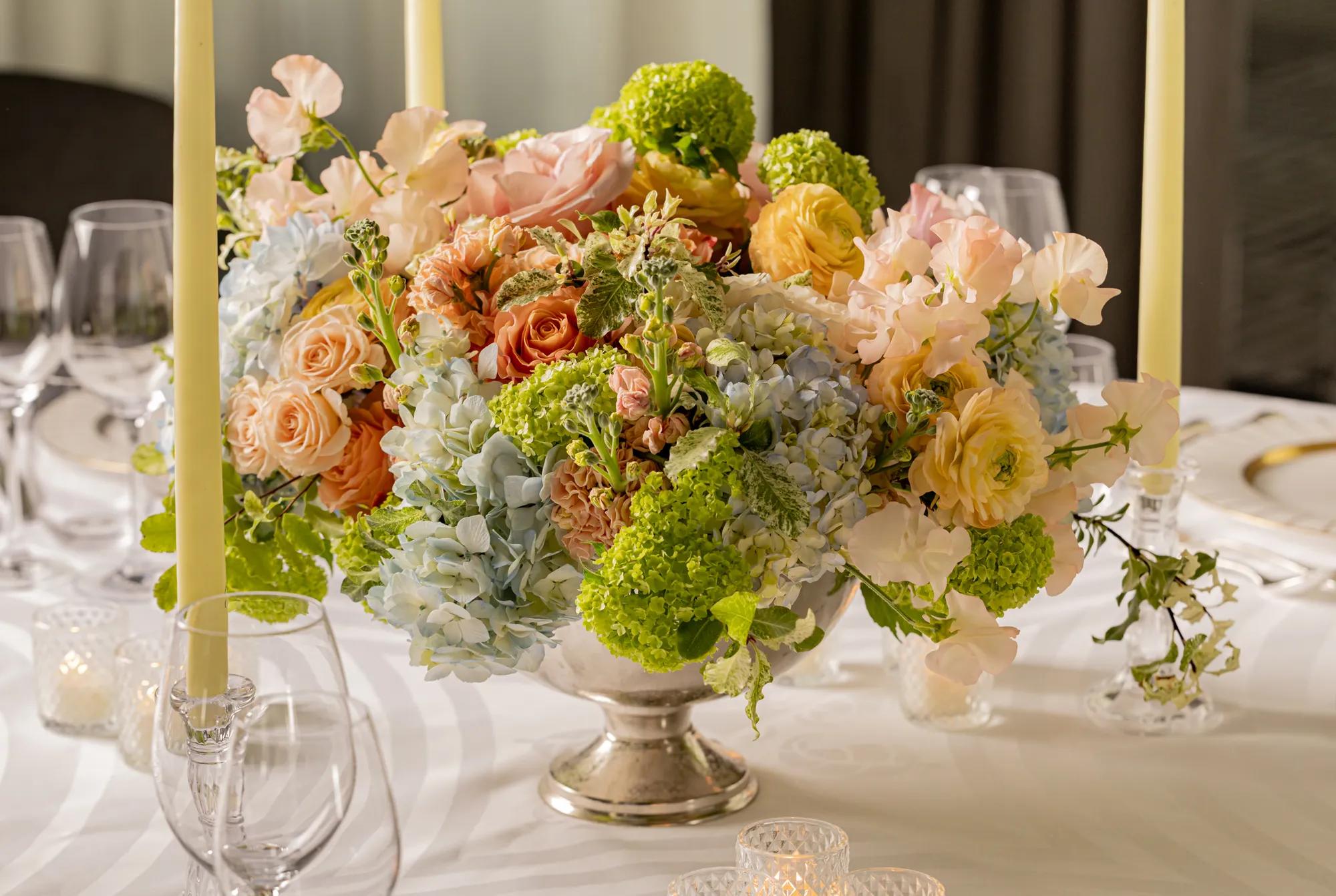 Pastel floral centrepiece with roses, hydrangeas and green blooms on a formally set dining table with candles.