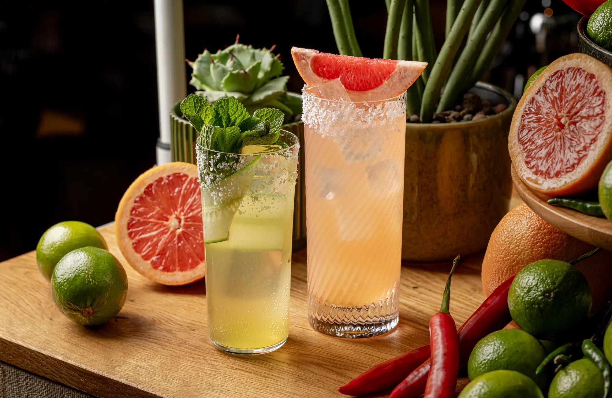 Two citrus cocktails garnished with mint and grapefruit sit among fresh limes, grapefruit and red chillies on a wooden display.